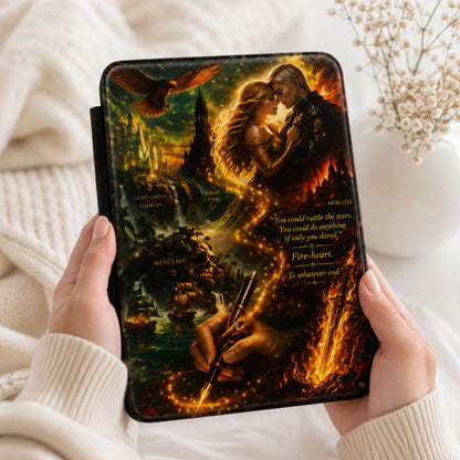 A person holds a hardcover book or notebook with a dramatic fantasy cover illustration featuring a glowing couple, a bird, and fiery scenes. The hands are covered in cream ribbed knit sleeves, and the background shows a cozy white blanket with small dried flowers.